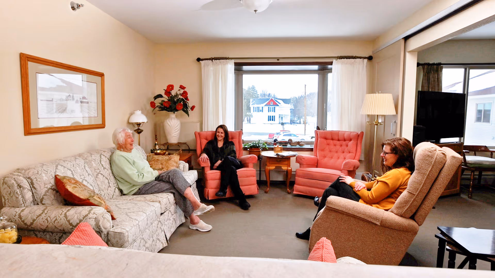 Three people sit and converse in a cozy senior-living common room furnished with sofas, armchairs, side tables and a large window.