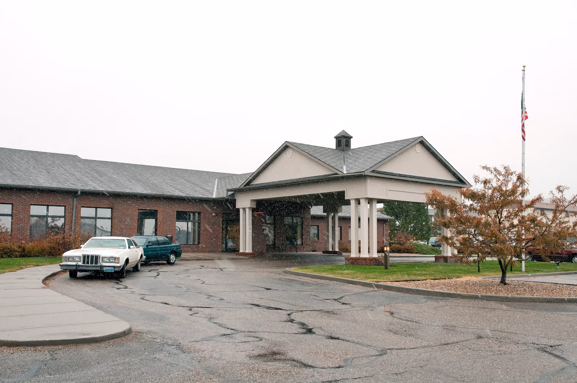 Exterior view of the Life Care Center of Casper building on a rainy day, showing a covered entrance with columns, a flagpole with an American flag, two parked cars, and some trees with autumn foliage.
