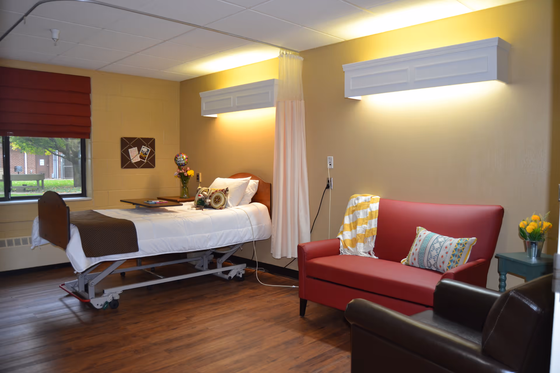 A well-lit room with a hospital-style bed made up with white linens and a brown blanket, positioned near a window with a red blind. Next to the bed is a small table with flowers and a balloon. The room also features a red loveseat with a colorful pillow and a yellow and white striped throw blanket, a dark brown armchair, and a small green side table with a vase of yellow flowers. The walls are painted beige and have two wall-mounted light fixtures above the bed and loveseat.