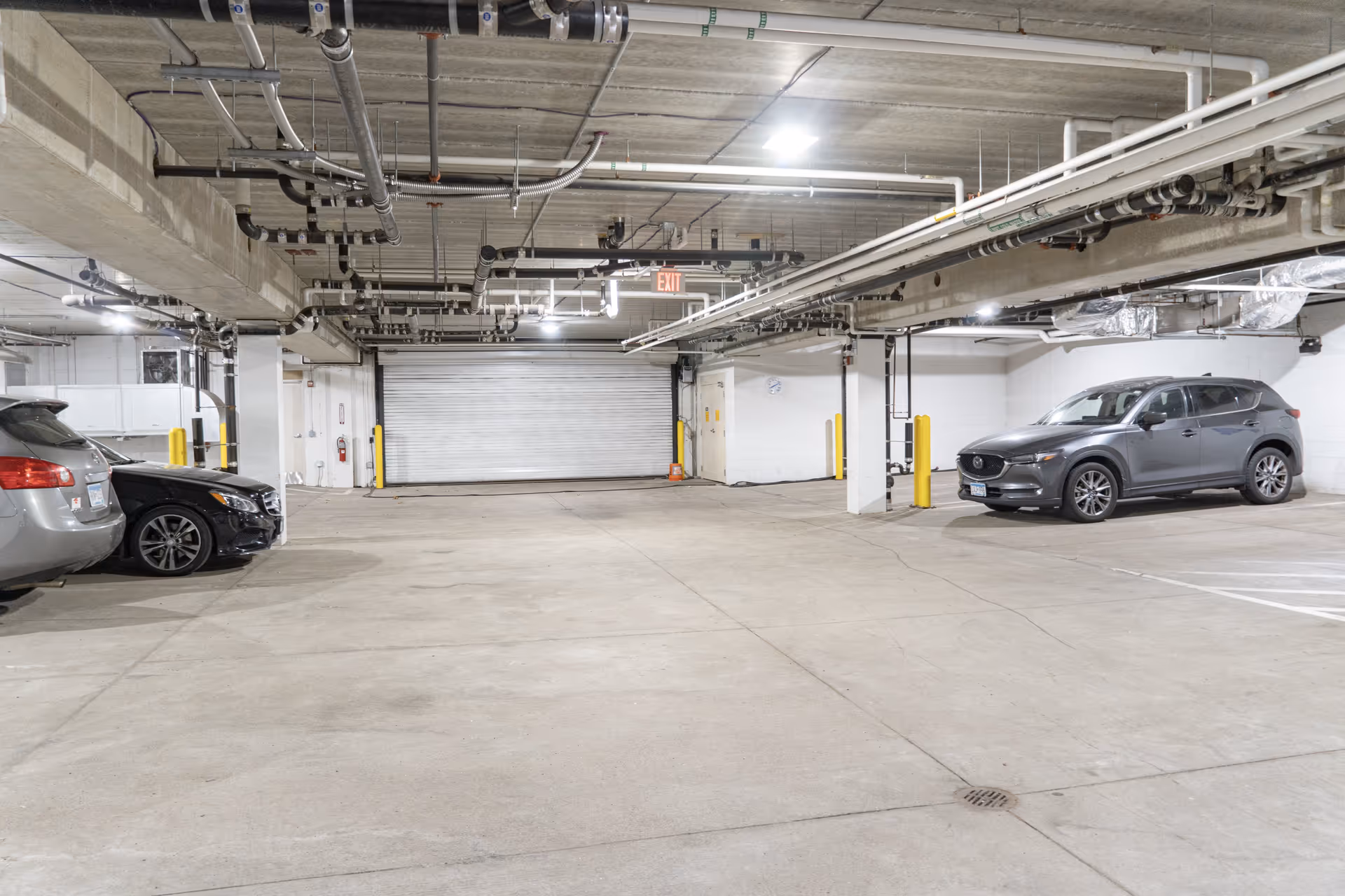 Indoor parking garage with concrete floors and ceiling, several parked cars, exposed pipes and lighting fixtures on the ceiling, and a closed rolling door at the far end.