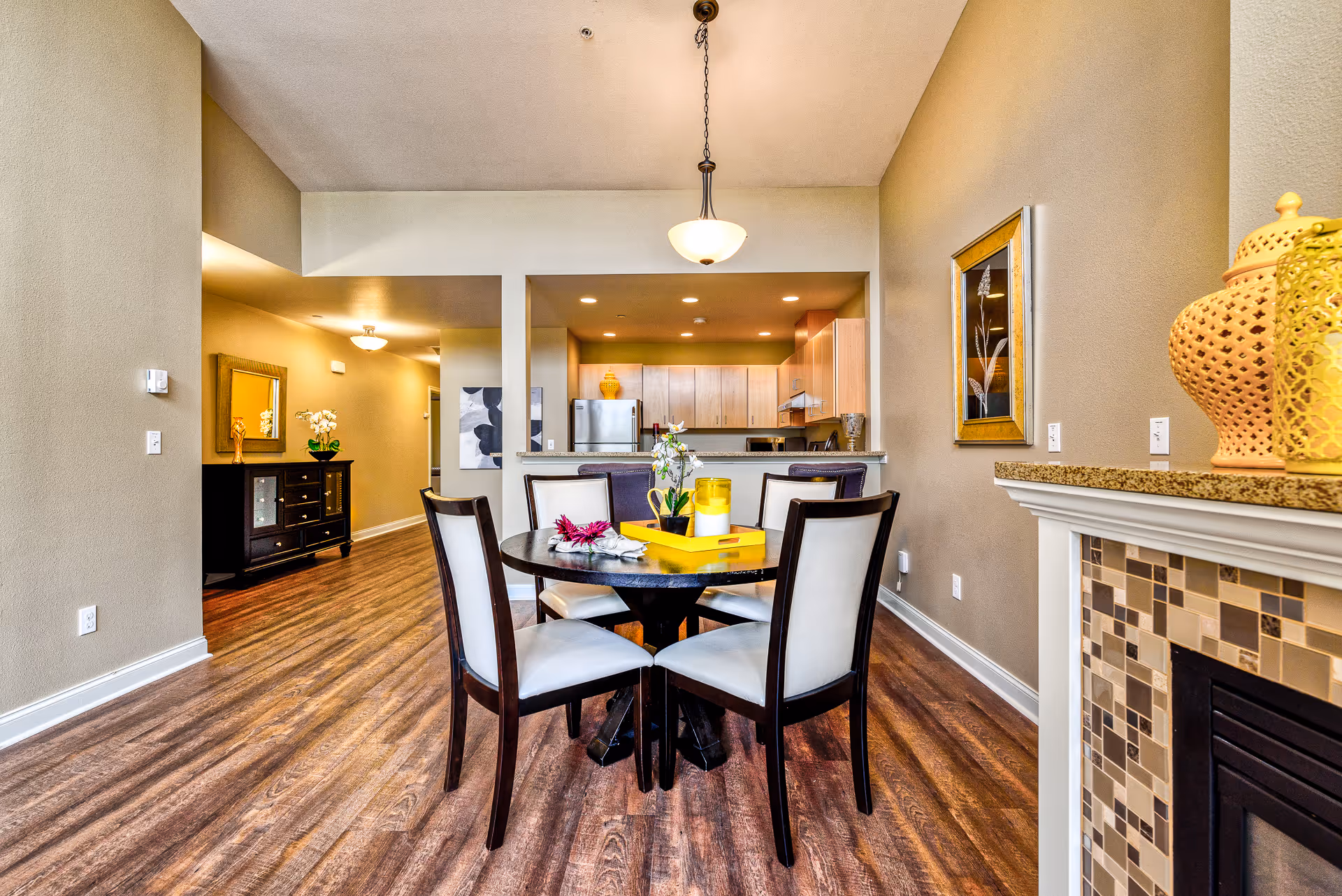 Interior view of a senior living facility dining area with a round table surrounded by six chairs. The table has a yellow tray with decorative items and flowers. In the background, there is a kitchen with wooden cabinets, a refrigerator, and a countertop with bar stools. The room has wooden flooring, beige walls, a hanging light fixture, and a fireplace with mosaic tile detailing on the right side.