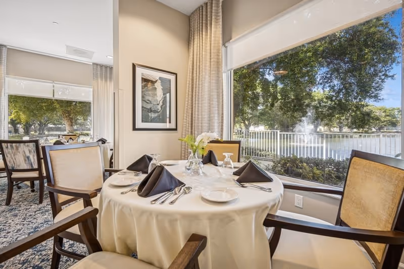 A dining area with a round table covered with a white tablecloth, set with plates, silverware, glasses, and black folded napkins. Four wooden chairs with beige cushions surround the table. Large windows provide a view of a lake with a fountain and trees outside. Light-colored curtains and a framed artwork decorate the interior.