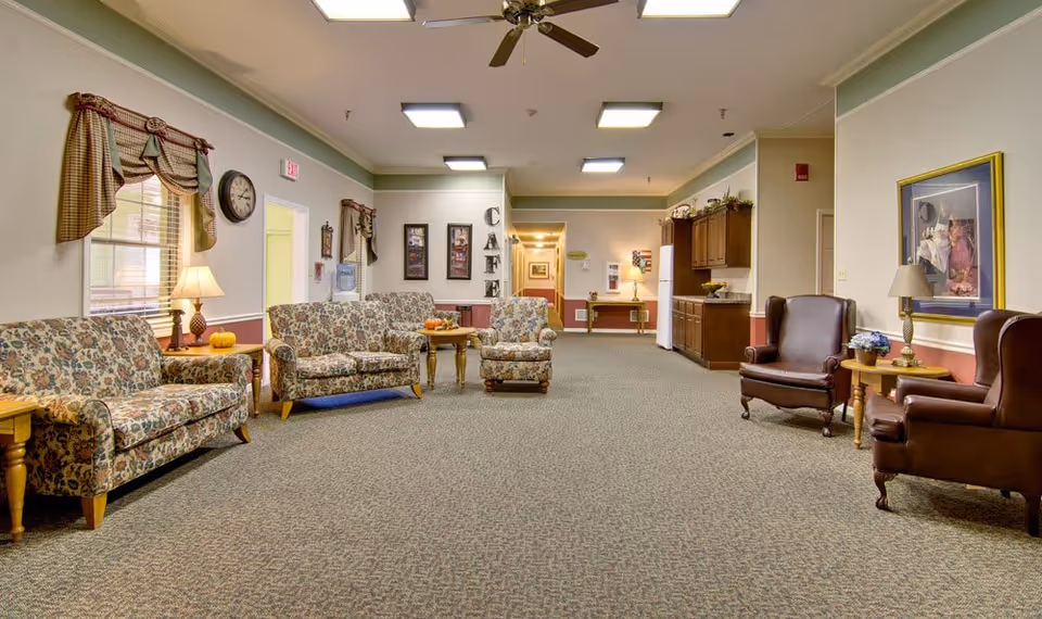A spacious senior living facility common area with floral patterned sofas and armchairs arranged around wooden side tables. The room has beige walls with a green trim, carpeted floor, ceiling fans, and fluorescent lighting. There is a kitchenette area with cabinets and a refrigerator on the right side, and a wall with the word 'CAFE' displayed vertically. Windows have plaid valances, and framed artwork and lamps decorate the space.