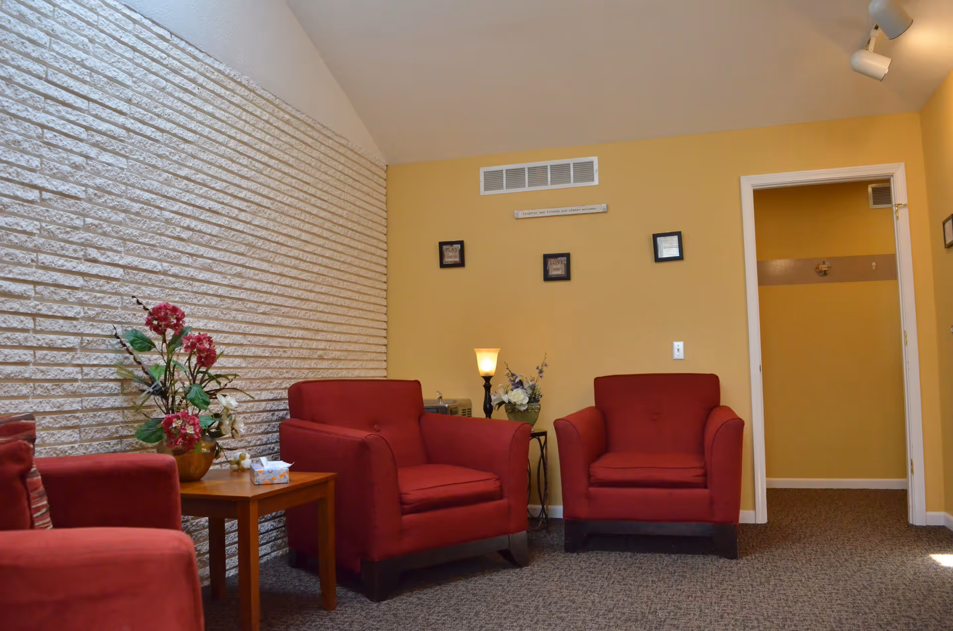 Small seating area with red armchairs, a side table, lamp and floral arrangement against a yellow wall and textured white brick accent.