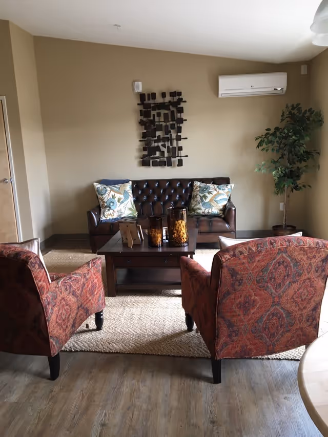 A cozy living room with a dark brown leather tufted sofa adorned with two floral patterned pillows. In front of the sofa is a wooden coffee table with decorative items including three amber-colored glass candle holders. Two upholstered armchairs with a red and orange patterned fabric face the sofa. The room has light beige walls, a wooden floor, a woven area rug, a tall green potted plant in the corner, and a white wall-mounted air conditioning unit above the sofa. A modern black metal wall art piece is hung above the sofa.