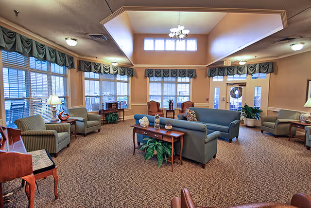 A spacious living room area with multiple seating options including sofas and armchairs arranged around a central coffee table. Large windows with green valances allow natural light to fill the room. The ceiling is high with a chandelier hanging in the center. There are plants and decorative items placed on tables throughout the room.