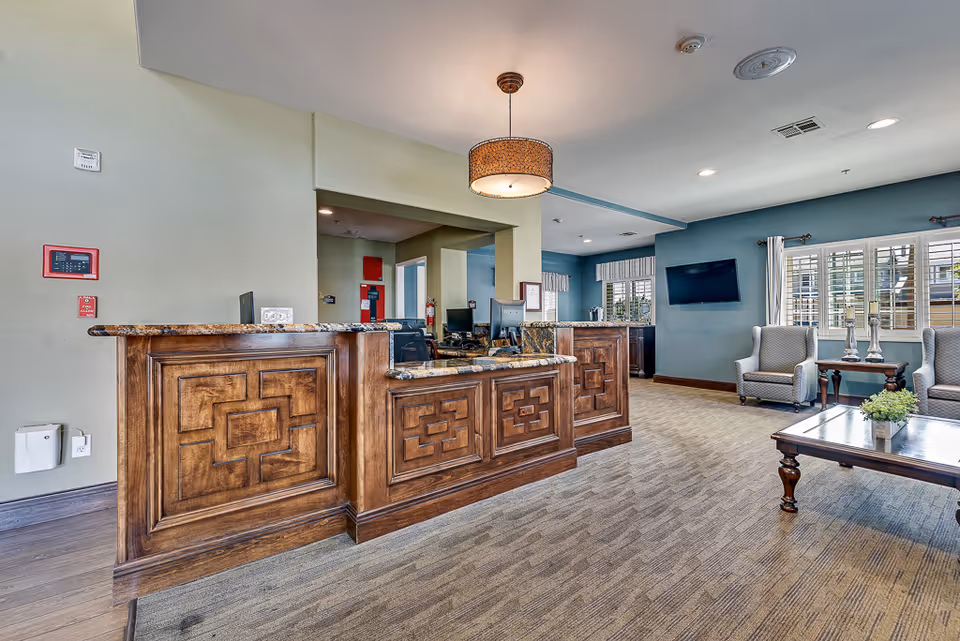 Reception desk and seating area in a senior living facility lobby with a wooden front desk, chairs, and a coffee table.