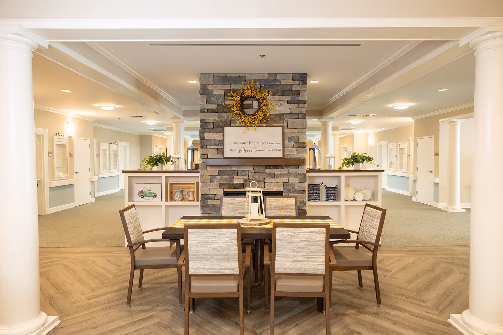 A cozy common area in a senior living facility featuring a wooden table with six cushioned chairs arranged around it. Behind the table is a stone fireplace decorated with a yellow floral wreath and a framed sign. The room has white columns, soft lighting, and built-in shelves with decorative items. The flooring is a combination of wood and carpet, and the space appears warm and inviting.