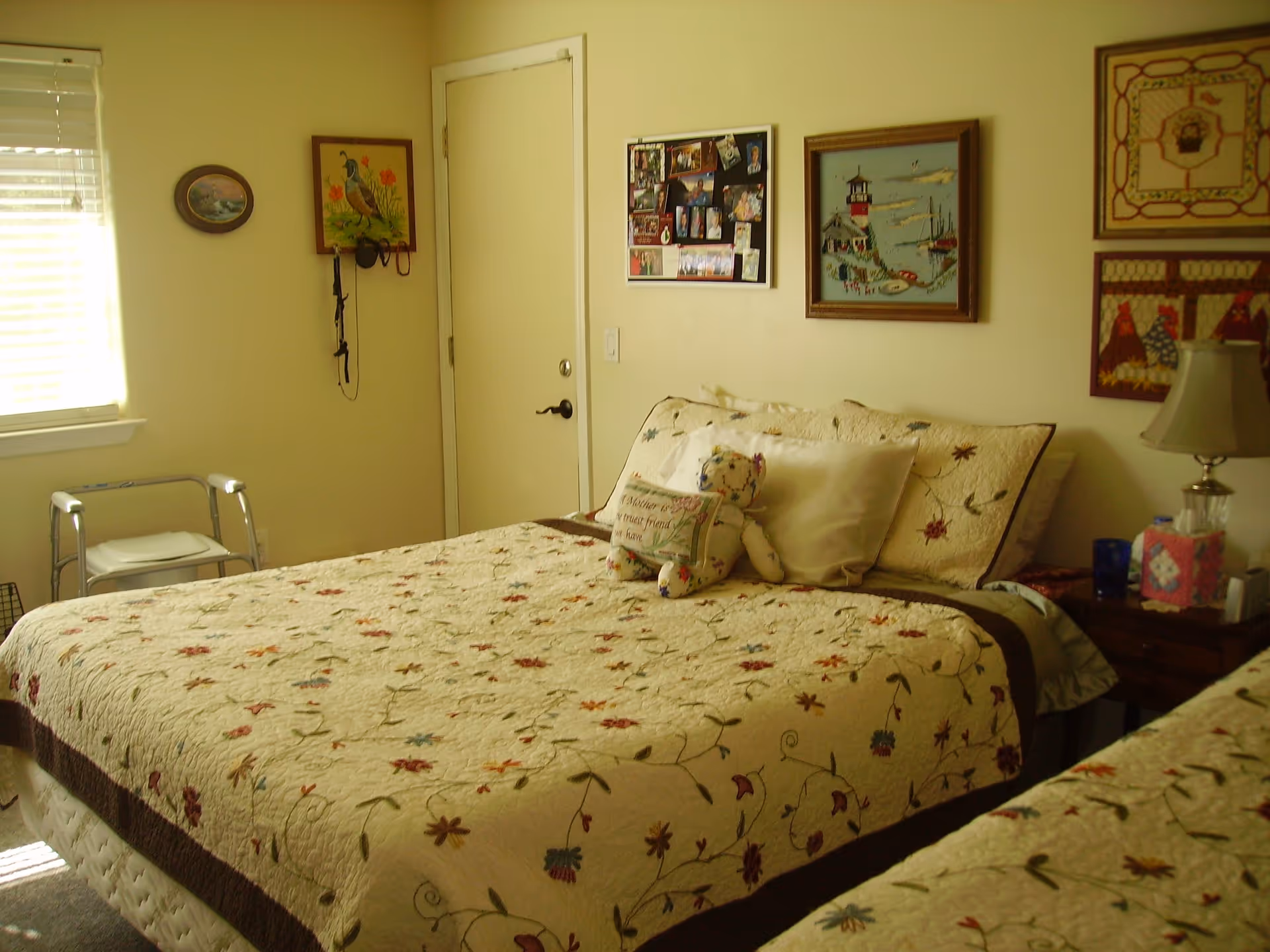 A cozy bedroom with a bed covered in a floral quilt, a teddy bear holding a pillow, a window with blinds, a walker near the window, and several framed pictures and artwork on the walls.