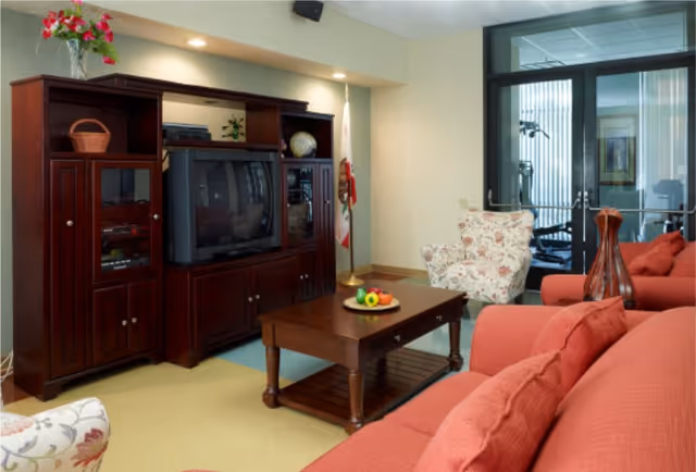 A cozy living room lounge with red sofas, a wooden entertainment center holding a TV, a coffee table with fruit, and glass doors to an adjoining room.