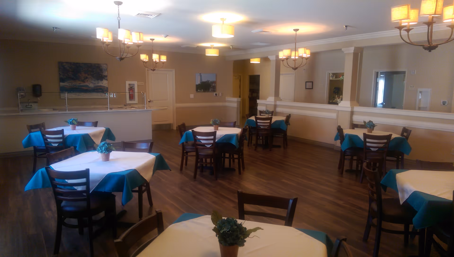 Interior view of a dining room with several tables covered with white and teal tablecloths, each table having a small potted plant centerpiece. The room has wooden flooring, beige walls, multiple chandeliers with warm lighting, and a counter area in the back with a painting on the wall.