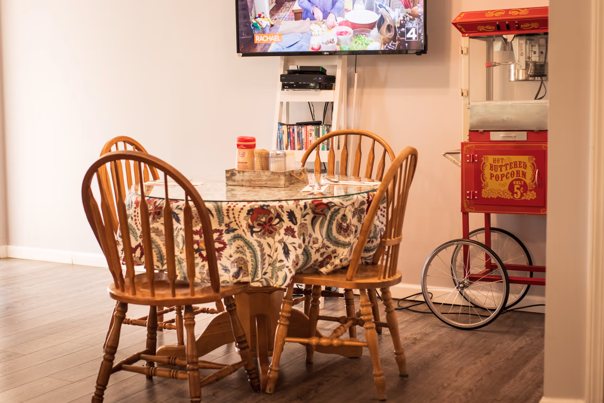 A cozy dining area with a round wooden table covered with a floral tablecloth and surrounded by four wooden chairs. On the table, there is a wooden tray holding condiments. In the background, a red vintage-style popcorn machine and a wall-mounted TV are visible.