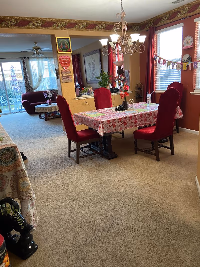 A dining room with a rectangular table covered with a heart-patterned tablecloth and four red upholstered chairs. A chandelier with multiple lights hangs above the table. The room has beige carpet and red curtains on the windows. There is a 'Happy Birthday' banner hanging on the window. In the background, a living room area with a maroon sofa and a round table with a lace tablecloth is visible, along with sliding glass doors leading outside.