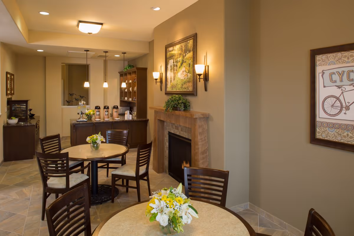 Cozy common dining area with round tables and chairs, a fireplace, coffee station, and floral centerpieces.