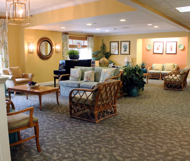 A spacious and well-lit living room area in an assisted living facility with comfortable wicker and cushioned seating, a wooden coffee table, potted plants, framed artwork on the walls, and large windows with curtains allowing natural light to enter.
