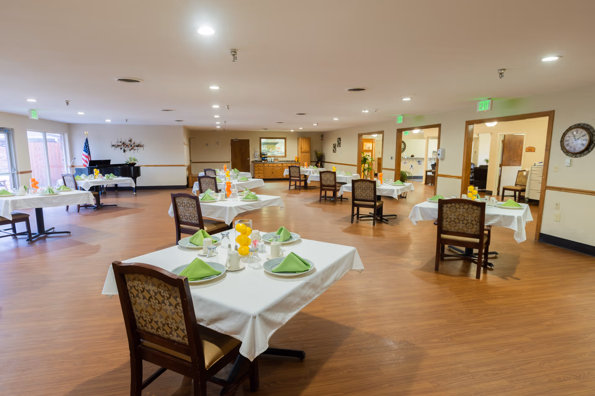 A spacious dining room with multiple tables covered in white tablecloths, each set with green folded napkins, plates, cups, and centerpieces featuring yellow lemons. The room has wooden flooring, beige walls with wooden trim, and several chairs with patterned upholstery. Large windows on the left side allow natural light in, and an American flag and a piano are visible in the background. There are exit signs above doorways on the right side of the room.