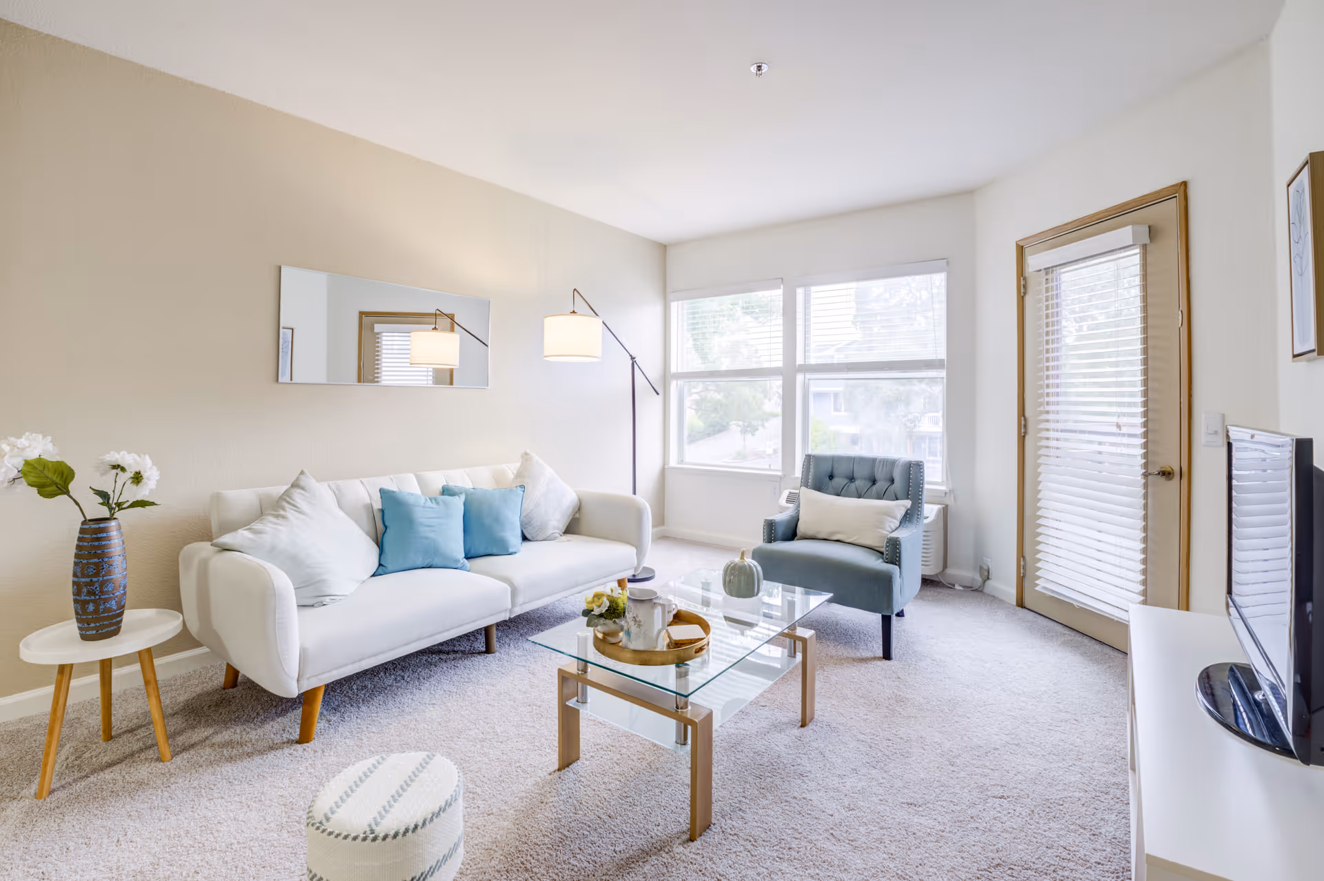 A bright and cozy living room with a white sofa adorned with blue and white pillows, a blue armchair with a white cushion, a glass coffee table with decorative items, a small round side table with a vase of flowers, a floor lamp, a wall mirror, large windows letting in natural light, and a TV on a white stand.