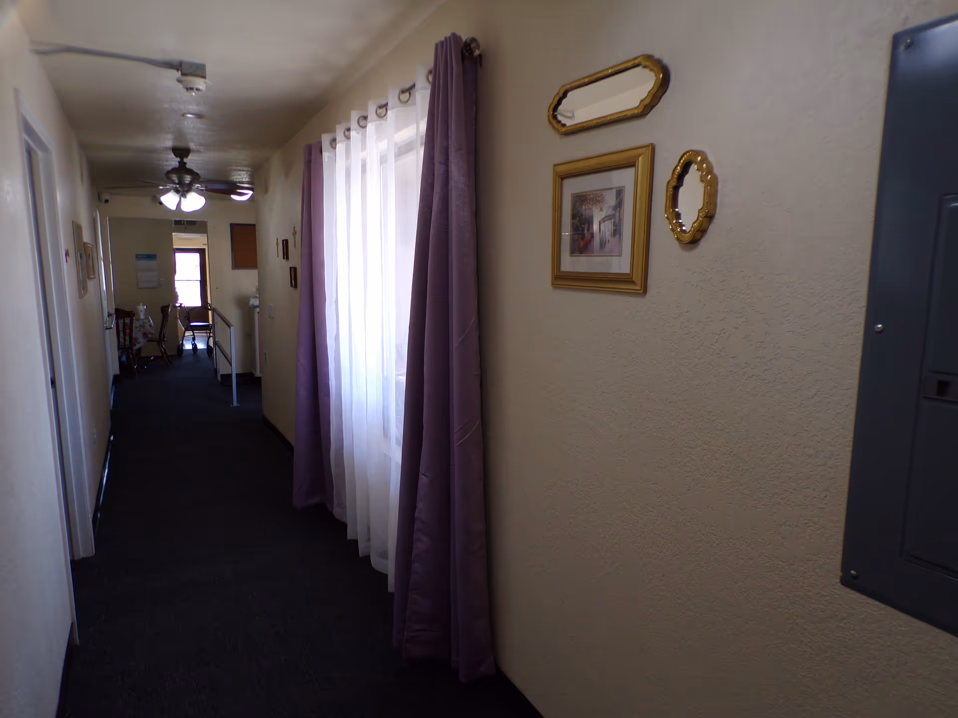 A narrow hallway with dark carpet flooring and cream-colored walls. On the right wall, there are three decorative items: two small mirrors with gold frames and a framed picture. A window with sheer white curtains and purple drapes is also on the right side. At the end of the hallway, there is a ceiling fan with lights, a chair, and a walker near a table.