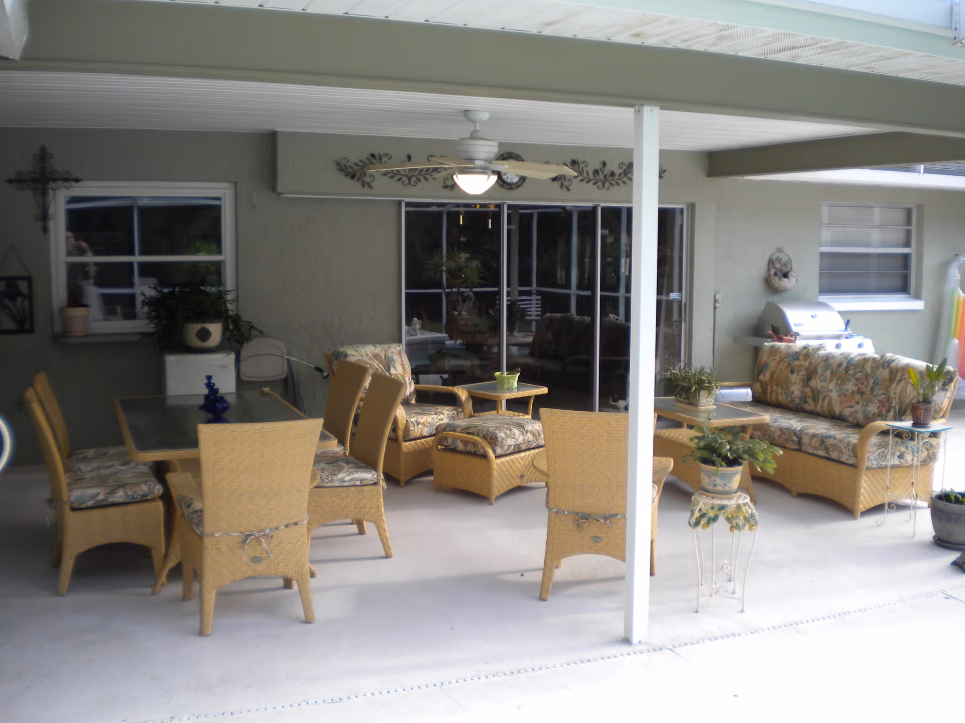 Covered outdoor patio area with wicker furniture including a dining table with six chairs, a cushioned sofa, two cushioned armchairs, and a small table. There are several potted plants, a ceiling fan with light, a wall-mounted clock, and a barbecue grill against the wall. Sliding glass doors lead to an interior space.