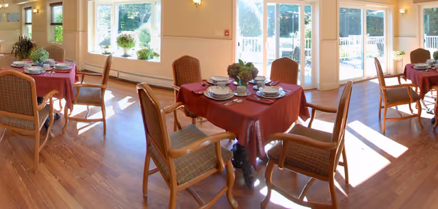 Sunlit dining room with tables covered in red tablecloths set for a meal, wooden chairs on hardwood floors, and large windows and glass doors overlooking greenery.