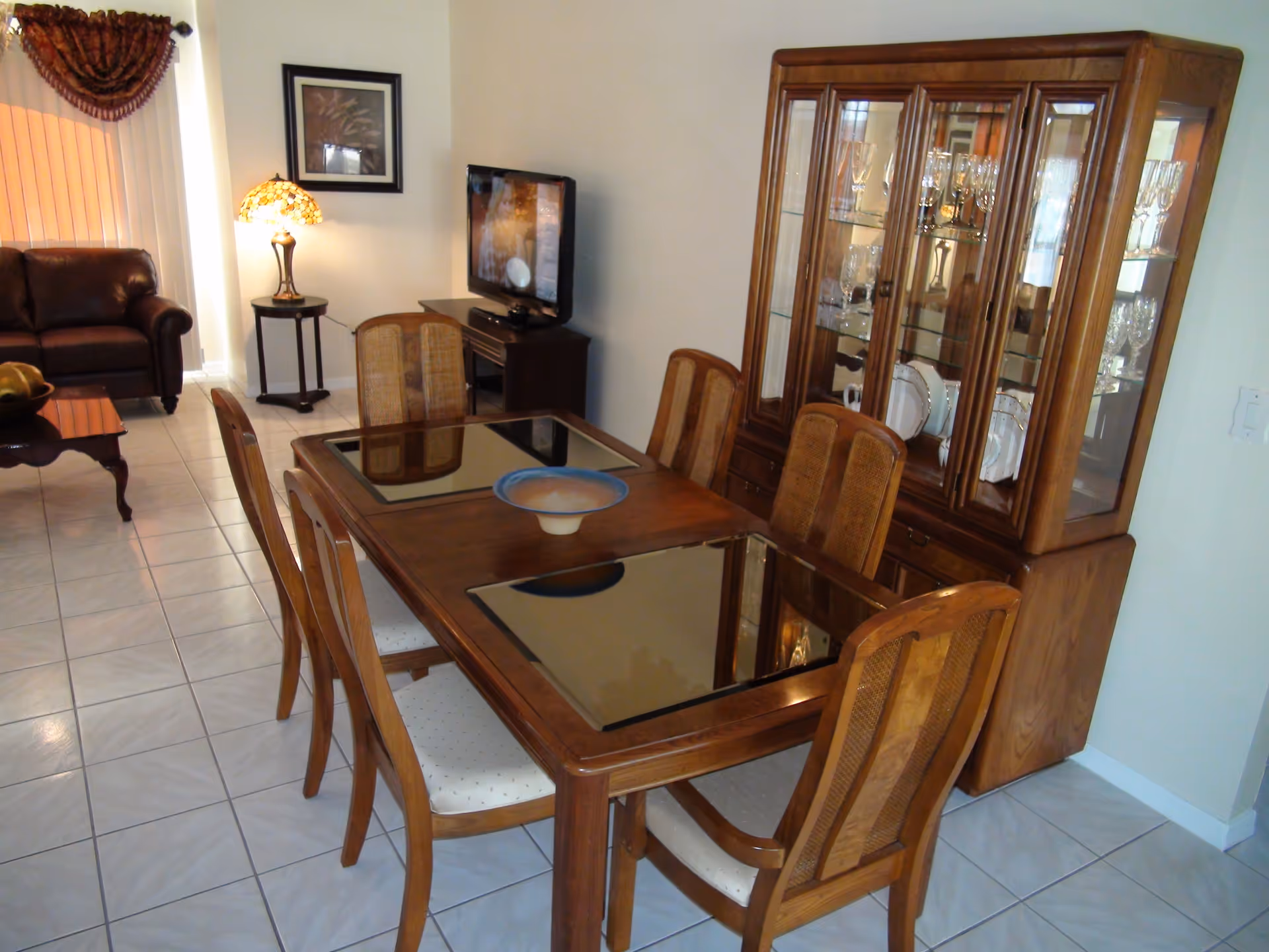 A dining area with a wooden table and six chairs, each chair having a cushioned seat. The table has two glass panels and a decorative bowl in the center. Behind the table is a wooden china cabinet displaying glassware and plates. To the left, there is a living area with a brown leather sofa, a side table with a decorative lamp, a wall picture, and a flat-screen TV on a stand. The floor is tiled with light-colored tiles.
