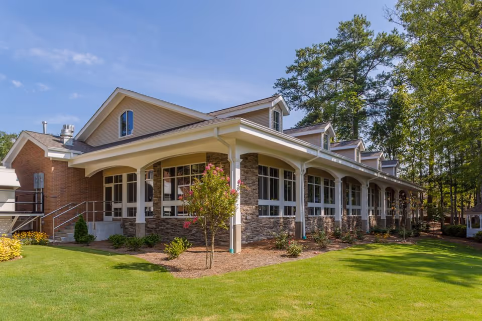 Exterior view of a single-story building with large windows, stone and brick facade, surrounded by green grass, small bushes, and trees under a clear blue sky.
