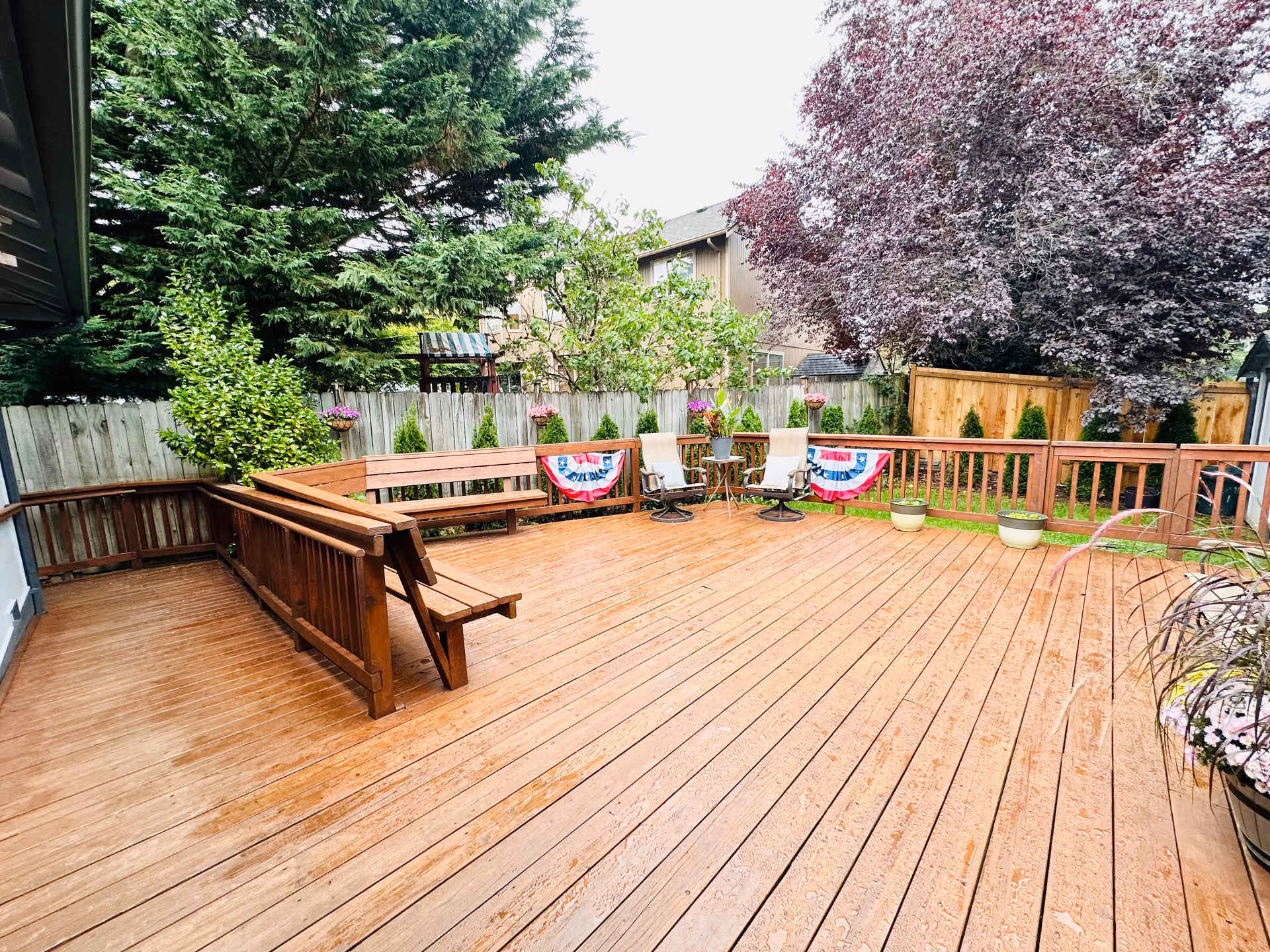 A spacious wooden outdoor deck with built-in benches and two chairs around a small table. The deck is decorated with patriotic bunting and surrounded by a wooden fence with various trees and plants in the background.