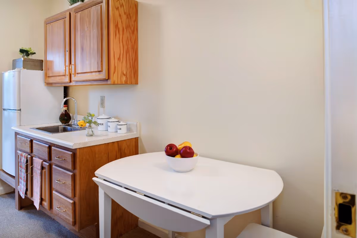 Small kitchenette with wooden cabinets, a sink and mini fridge next to a white drop-leaf table holding a bowl of fruit.