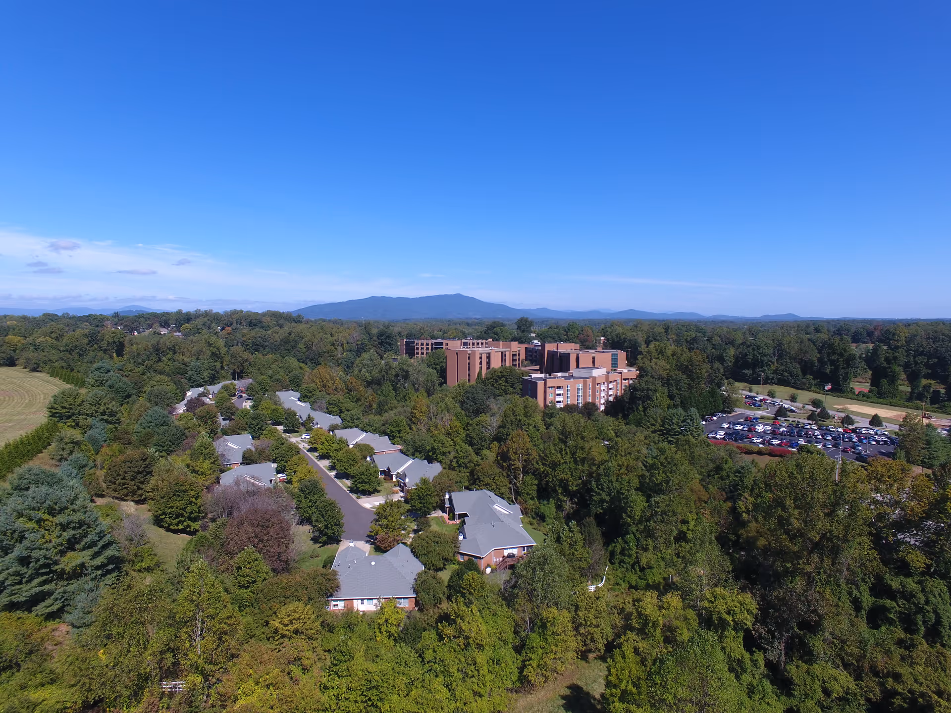 Aerial view of Westminster Canterbury facility surrounded by dense green trees with multiple buildings, a parking lot filled with cars, and mountains visible in the distance under a clear blue sky.
