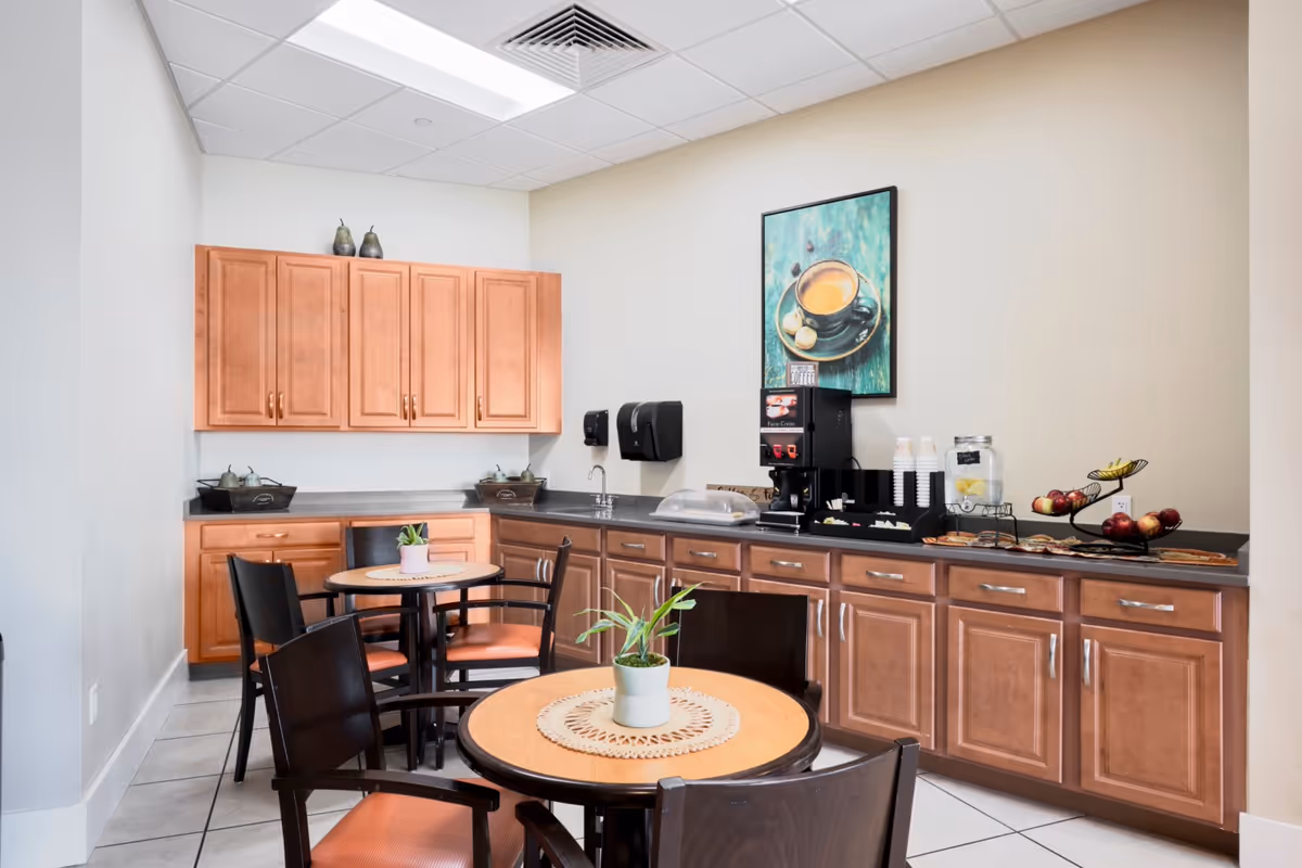 A small dining area with two round wooden tables, each surrounded by four dark wooden chairs with orange cushions. The room has light-colored walls and a tiled floor. Along one wall, there are wooden cabinets with a countertop holding a coffee machine, disposable cups, a water dispenser with lemon slices, and a tiered fruit basket with apples and bananas. Above the countertop is a framed picture of a cup of coffee on a teal background.
