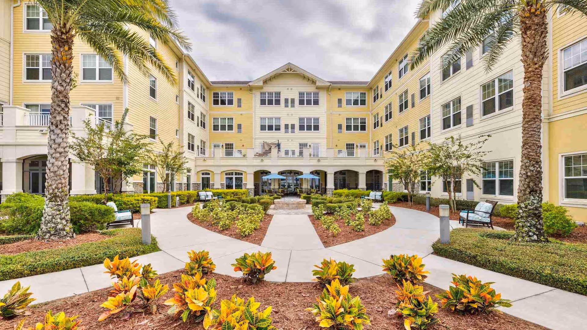 Yellow four-story senior living building surrounding a landscaped courtyard with palm trees, walking paths, benches, and planted beds.