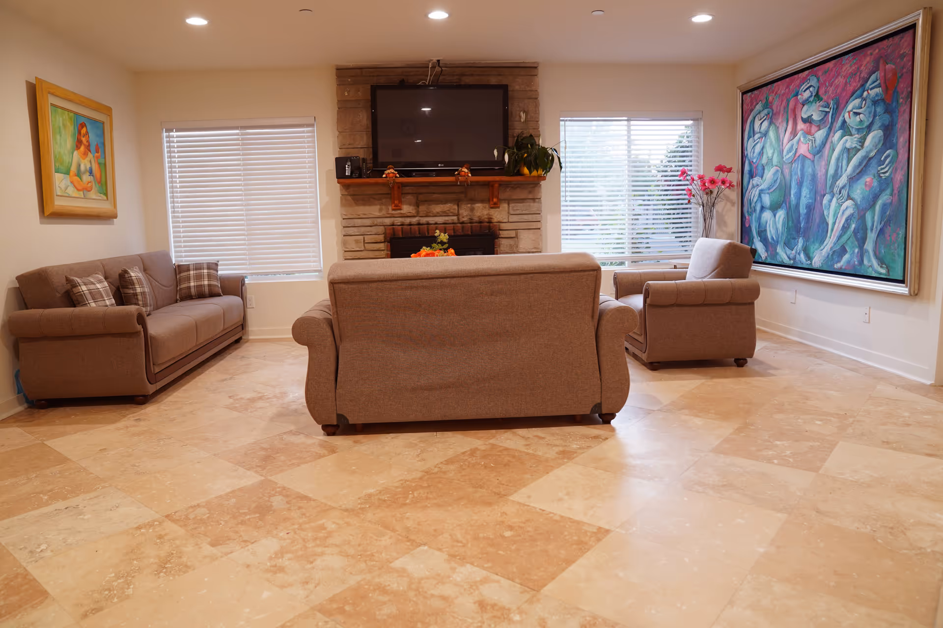 Living room with three beige sofas facing a fireplace and wall-mounted TV, tiled floor and large paintings on the walls.