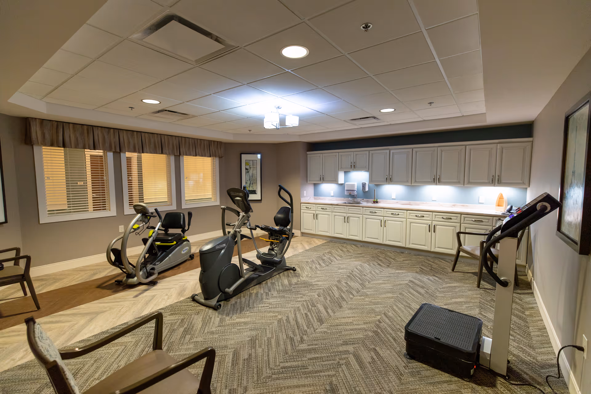 A fitness room with exercise equipment including two stationary bikes and a treadmill. The room has beige walls, a patterned carpet, overhead lighting, and a row of white cabinets along one wall. There are also a few chairs placed around the room and windows with blinds.