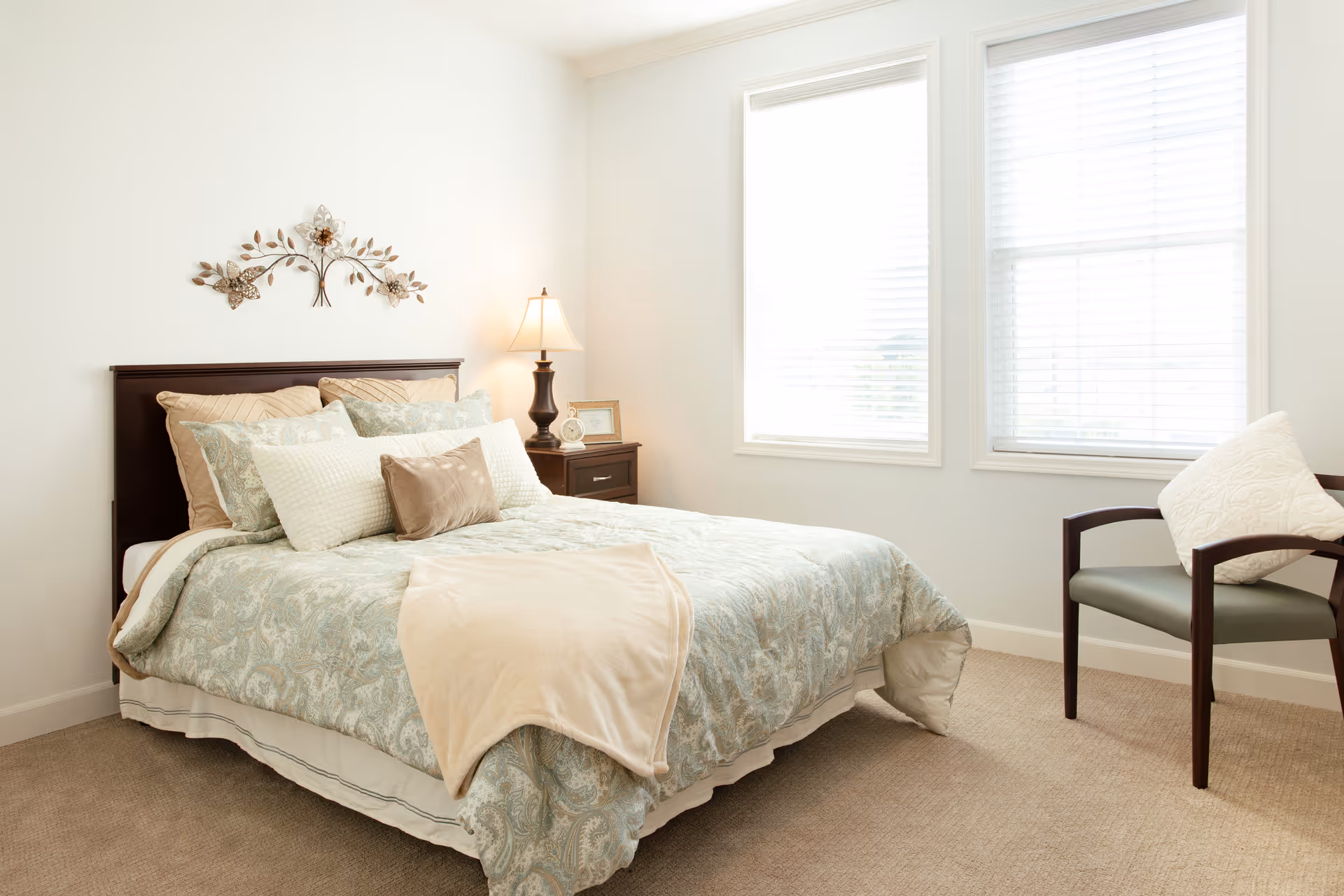 A bright bedroom with a neatly made bed featuring patterned bedding and multiple pillows. There is a wooden headboard with a decorative metal wall art above it. A nightstand with a lamp, a small clock, and a framed picture is beside the bed. Two large windows with white blinds allow natural light to fill the room. A cushioned chair with a white pillow is positioned near the windows on a beige carpeted floor.