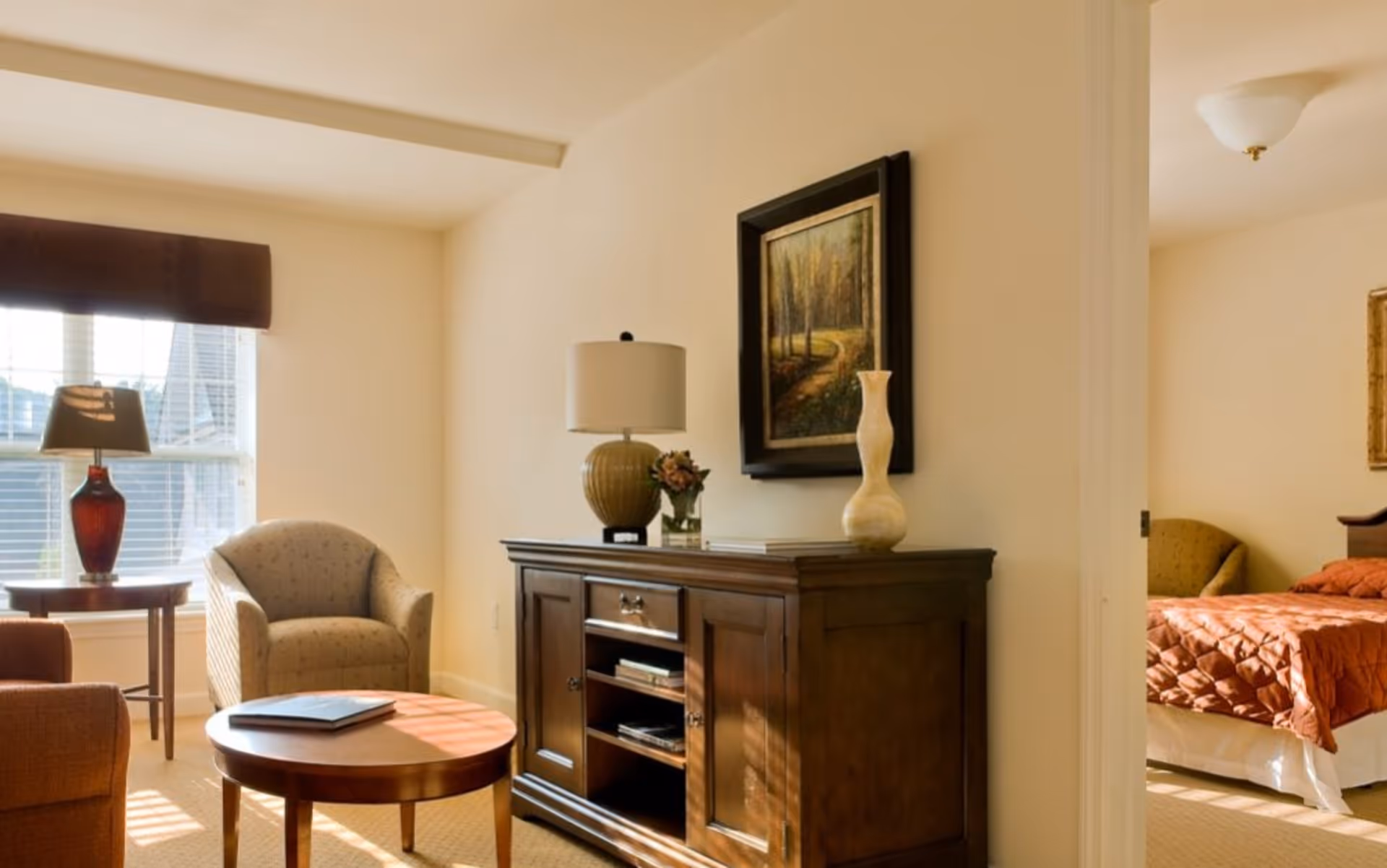 A cozy living room area with a round wooden coffee table, an armchair, a side table with a lamp, and a wooden cabinet with decorative items and a framed painting above it. Through an open doorway, a bedroom with a bed covered in a rust-colored quilt and an upholstered chair is visible.