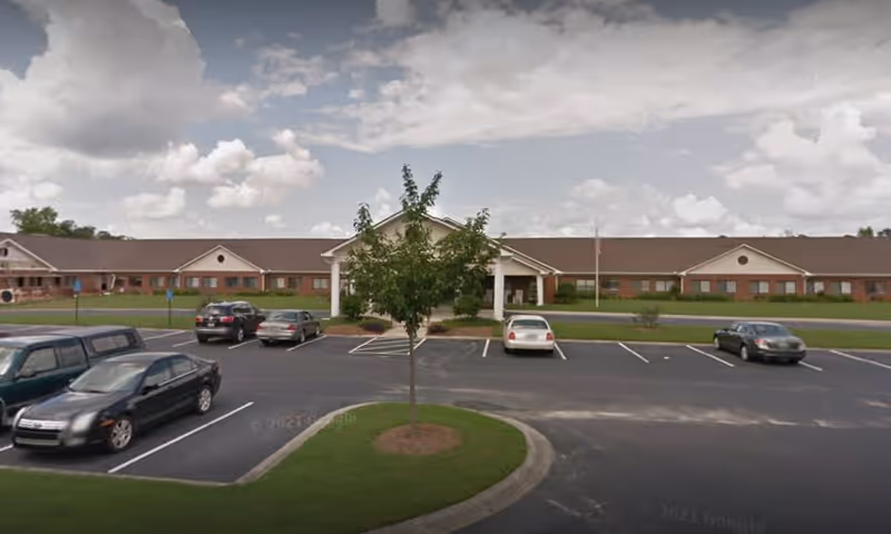 Front exterior view of a single-story senior living facility building with a brown roof and brick facade. There is a covered entrance in the center, a small tree in a landscaped island in the parking lot, and several parked cars. The sky is partly cloudy.