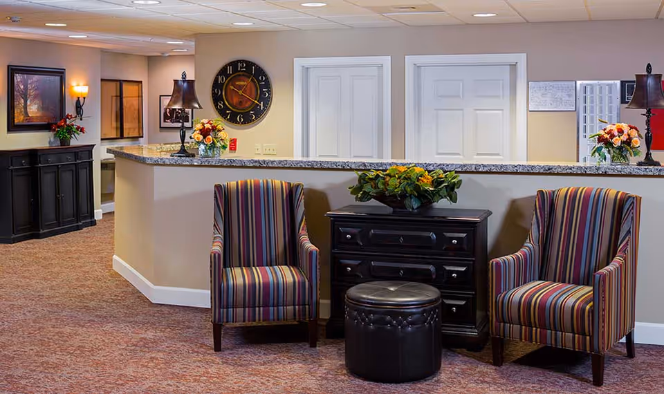 A cozy seating area in a memory care community featuring two striped armchairs with a small black ottoman between them. Behind the chairs is a dark wooden chest with a green plant on top. The background shows a granite countertop with two lamps and flower arrangements, a large wall clock, framed artwork, and two white doors.