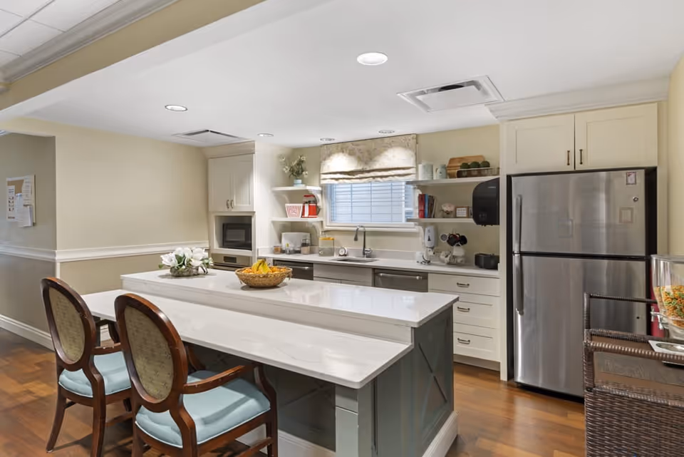 A clean and well-lit kitchen area with a white countertop island featuring a basket of bananas and a flower arrangement. Two wooden chairs with light blue cushions are placed at the island. The kitchen has white cabinets, a stainless steel refrigerator, a microwave, a sink under a window with a floral valance, and various kitchen items on shelves and counters.