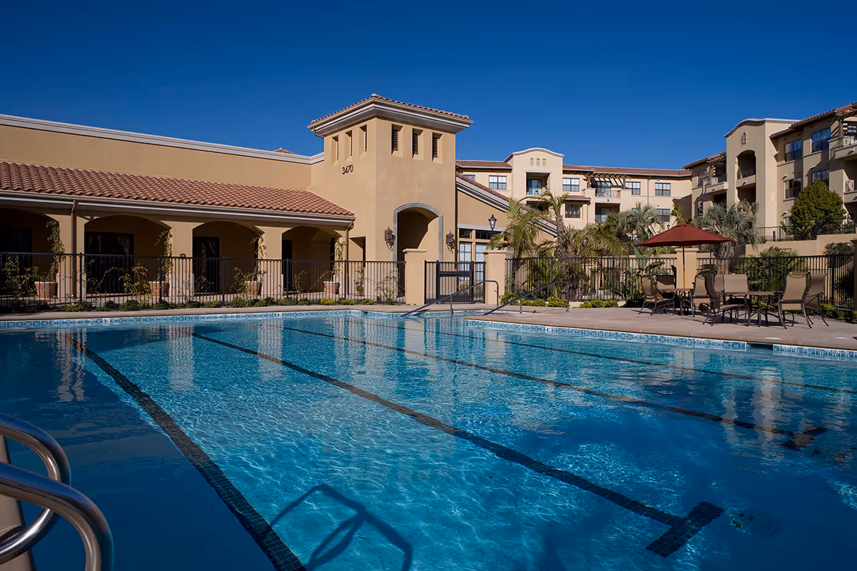 Outdoor swimming pool with clear blue water in front of a beige multi-story building with red tile roof and balconies. There are poolside chairs and tables with umbrellas, palm trees, and a clear blue sky.