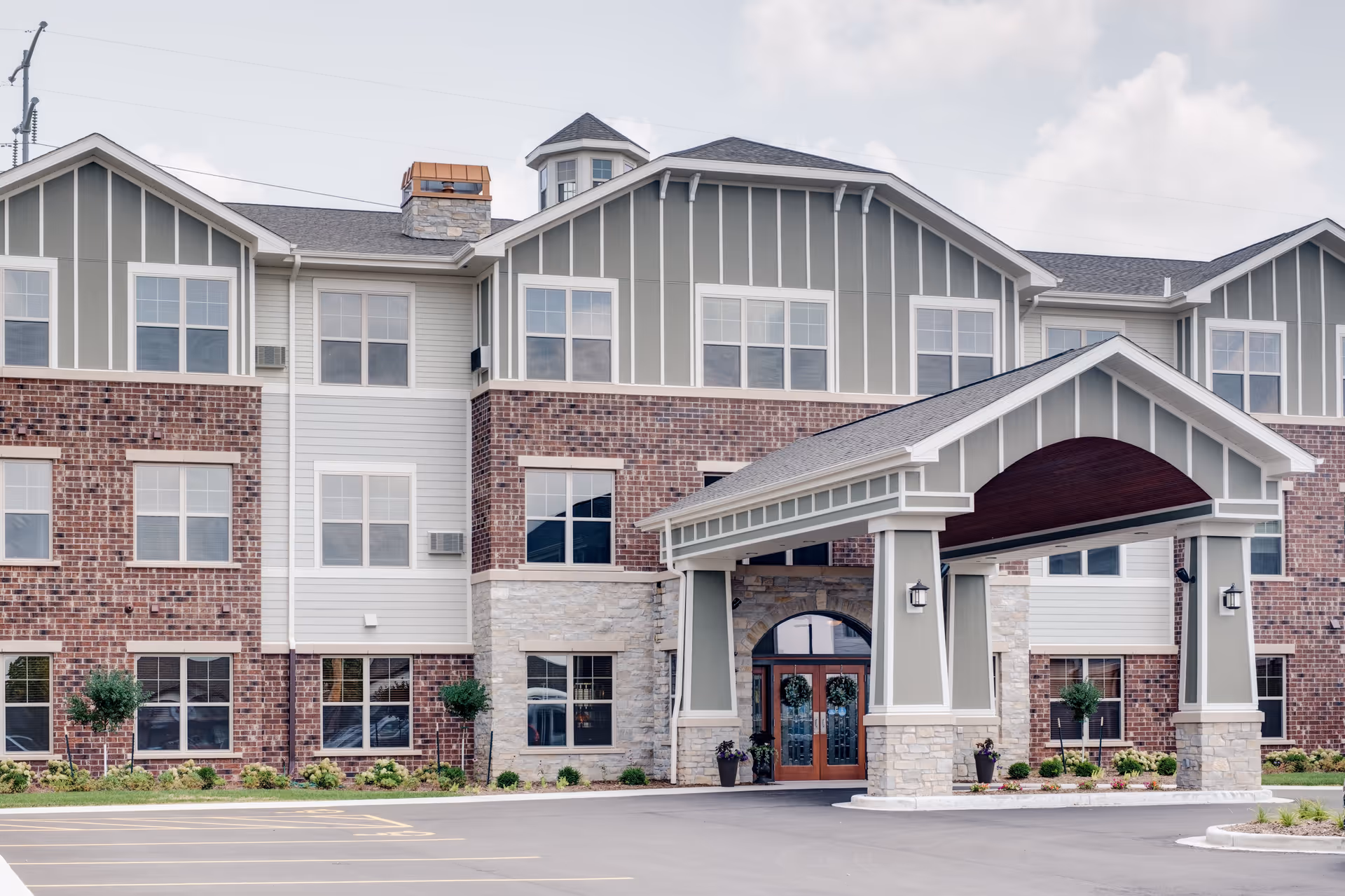 Exterior view of a three-story senior living facility building with a covered entrance, brick and siding facade, multiple windows, and landscaped greenery in front.