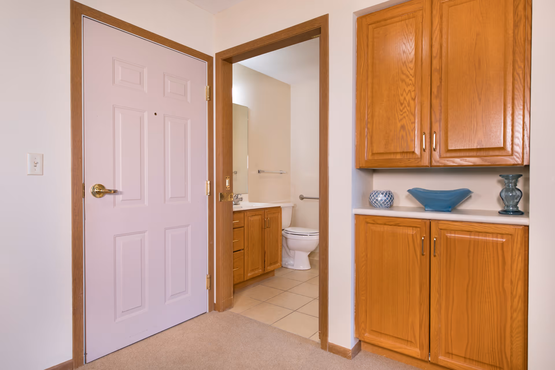 Entryway with a closed light-colored front door, an open doorway to a bathroom showing a toilet and vanity, and wooden built-in cabinets with decorative bowls.