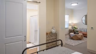 View from a bedroom into a bathroom and a living room area. The bedroom has a metal bed frame, the bathroom has a shower with a curtain, and the living room features a round mirror, a striped sofa, a wooden coffee table, and a patterned rug.