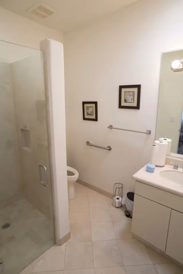 A clean bathroom with a glass-enclosed shower on the left, a toilet next to the shower, two framed pictures on the wall above a towel rack and grab bar, and a white vanity with a sink, paper towels, and a soap dispenser on the right.