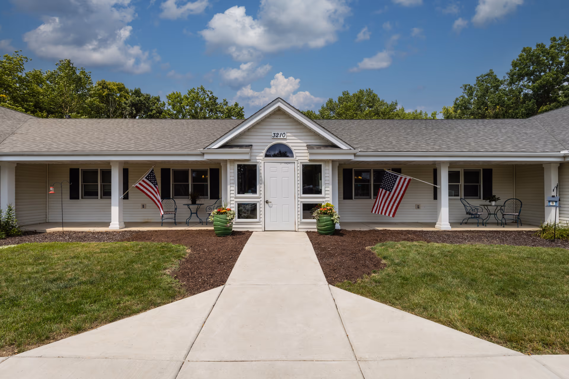 Front exterior view of a single-story building with a central white door, two American flags on either side, green potted plants, outdoor seating with tables and chairs, and a well-maintained lawn under a partly cloudy sky.
