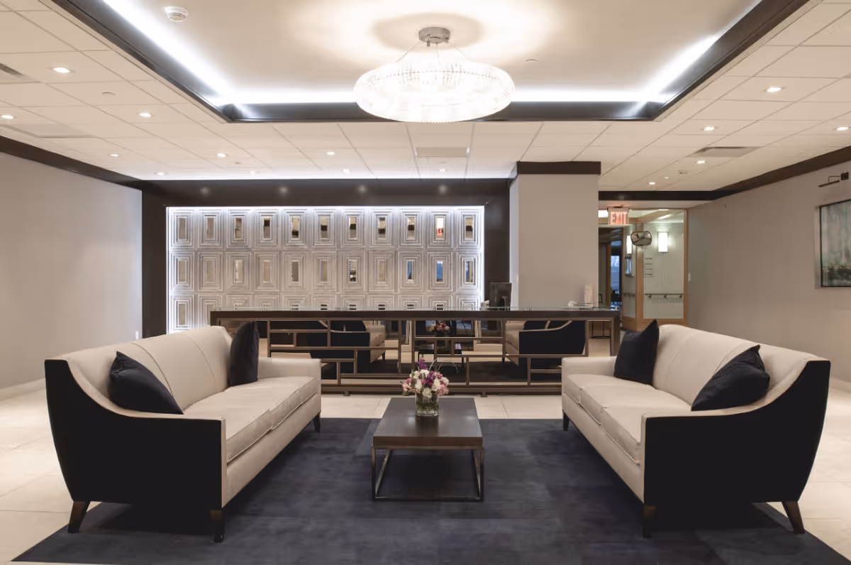 A modern and elegant lounge area with two beige sofas facing each other, each with black armrests and black cushions. Between the sofas is a dark wooden coffee table with a small flower arrangement. The floor is covered with a dark rug, and the ceiling features recessed lighting and a large decorative chandelier. The back wall has a geometric pattern with illuminated rectangular cutouts, and there is a mirror reflecting the room. The space has a calm and welcoming atmosphere.