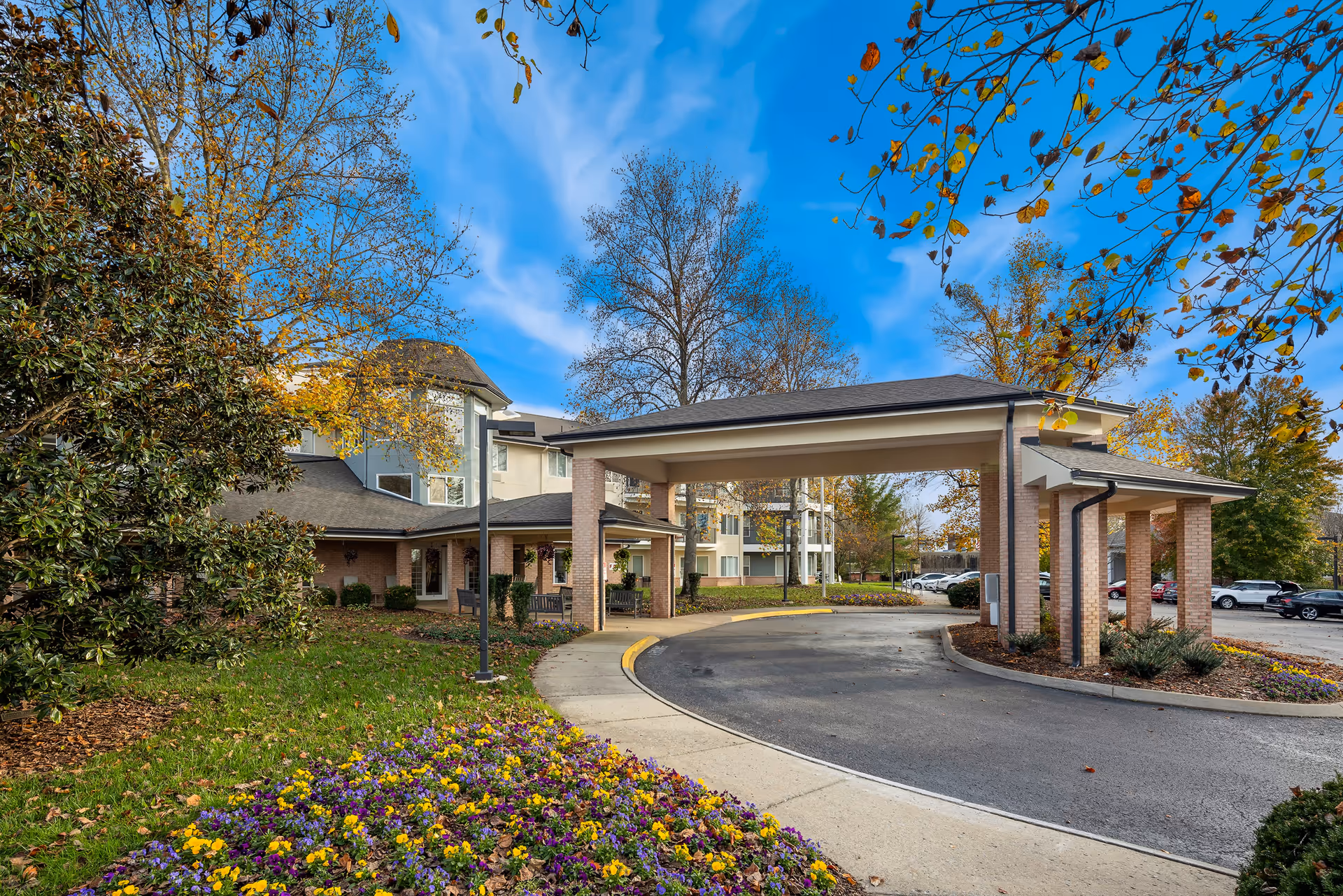 Entrance area of Manor at Steeplechase senior living community showing a covered driveway with brick pillars, surrounded by landscaped gardens with colorful flowers and trees with autumn leaves under a blue sky.