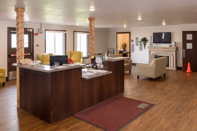 Interior view of a senior living facility reception area with a wooden front desk, office supplies, and seating including armchairs. The room has wooden flooring, a fireplace with a TV mounted above it, and a door leading to another room. Two wooden pillars wrapped with string lights are visible near the desk.