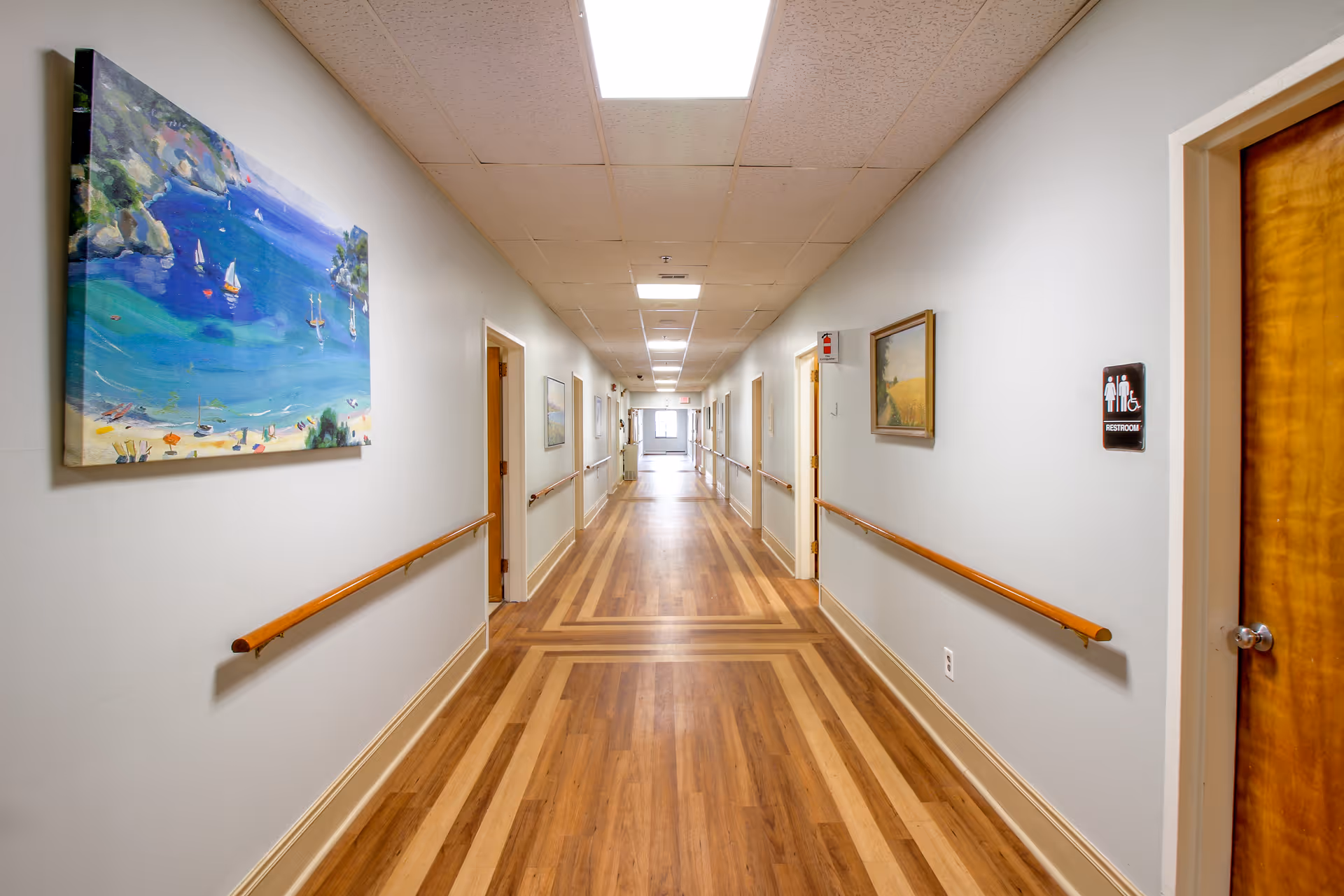 A long, well-lit hallway in a senior living community with wooden floors and handrails on both sides. The walls are decorated with paintings, and there are several doors along the corridor. A restroom sign is visible on the right side near a wooden door.