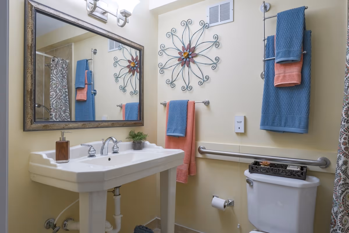 A clean and well-lit bathroom featuring a white pedestal sink with a silver faucet, a large rectangular mirror with a decorative frame above the sink, and a toilet with a decorative tray on top. The walls are light yellow, adorned with a metal wall art piece shaped like a flower. There are blue and coral towels hanging on silver towel racks, a patterned shower curtain partially visible, and a grab bar installed next to the toilet for accessibility.