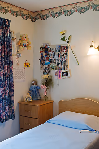 A cozy bedroom corner in a senior living facility with a single bed covered in a light blue blanket. Next to the bed is a wooden nightstand with three drawers, holding a doll in a blue dress, a small framed photo, and a vase with flowers. The wall above the nightstand is decorated with a collage of photos, a single yellow flower, and some hanging stuffed animals. Floral curtains cover the window, and a wall-mounted light fixture illuminates the area. The room has a floral wallpaper border near the ceiling.